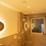 Modernes Badezimmer mit warmweißer Beleuchtung, Deckenspots und offener Dusche in stimmungsvoller Abendszene.