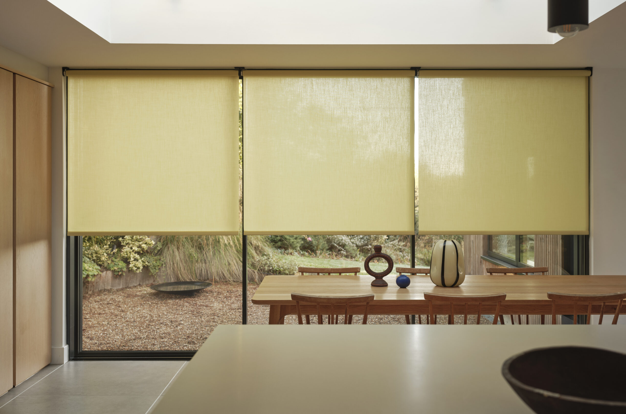 Essbereich mit großflächiger Fensterfront und textiler Beschattung durch elegante Rollos.
